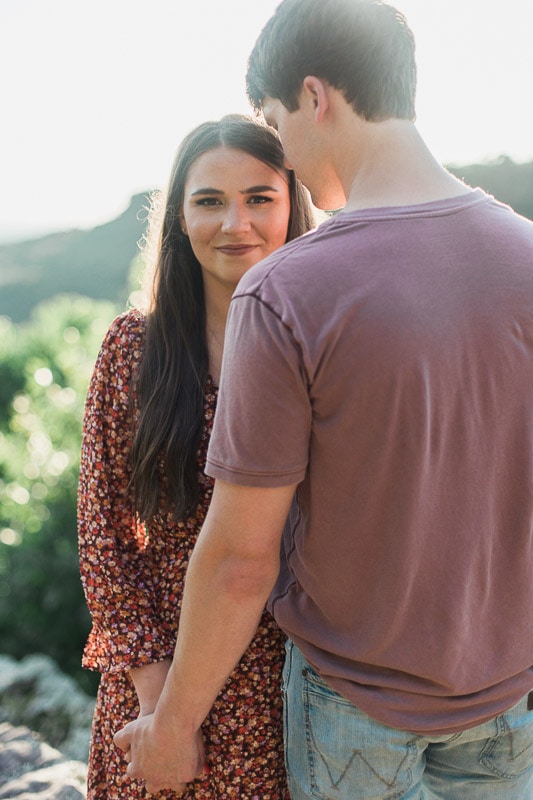 Girl looking at camera with fiancé's back to camera standing on a mountain. Photo by Arkansas Wedding Collection.