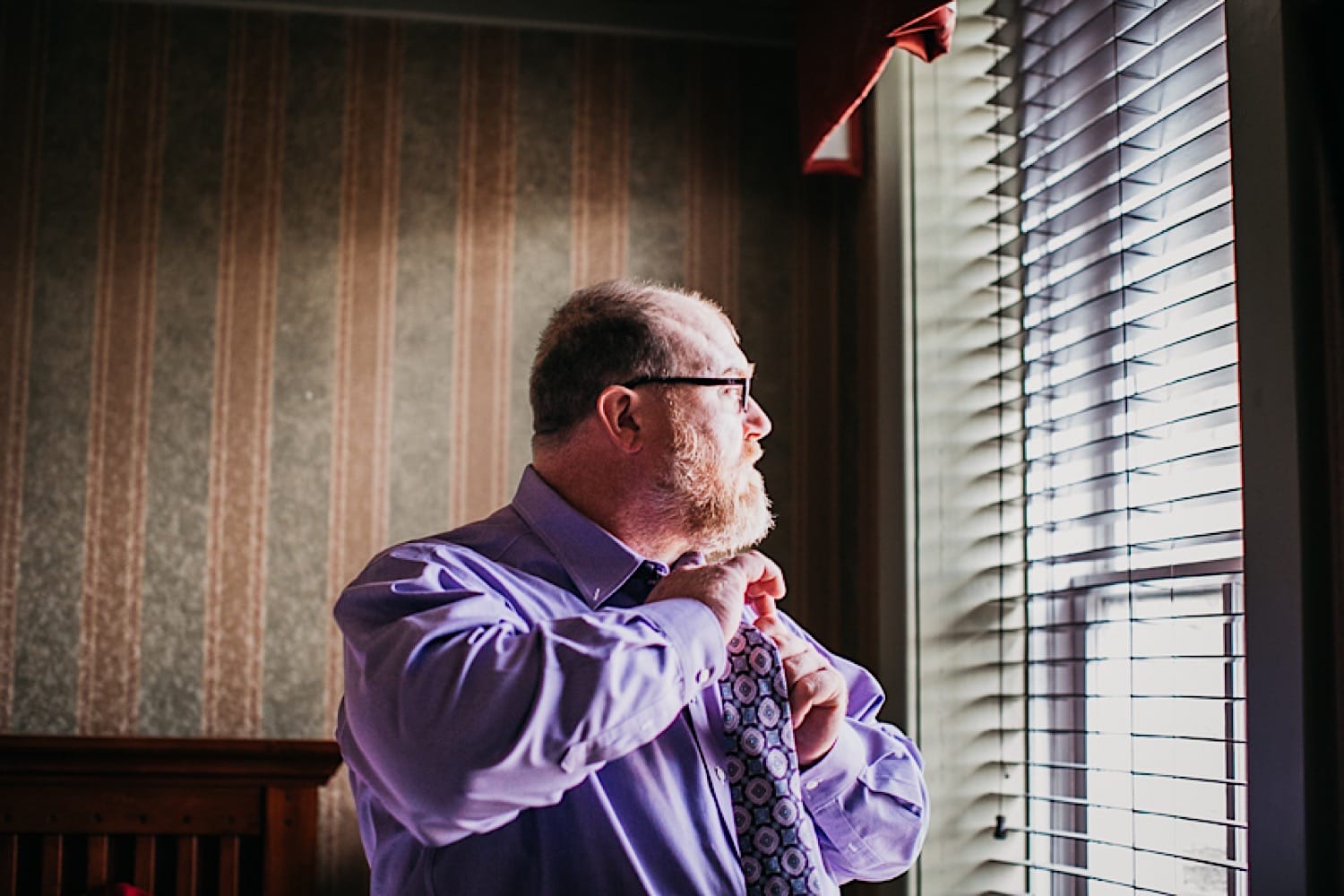 Groom putting on tie in groom’s quarters at Crescent Hotel while looking out window