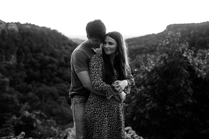 Black and white photo of couple holding each other while standing on overlook on a mountain at Petit Jean State Park. Photo by Arkansas Wedding Collection.